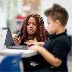 teacher kneels by desk of second grader who is looking at a laptop